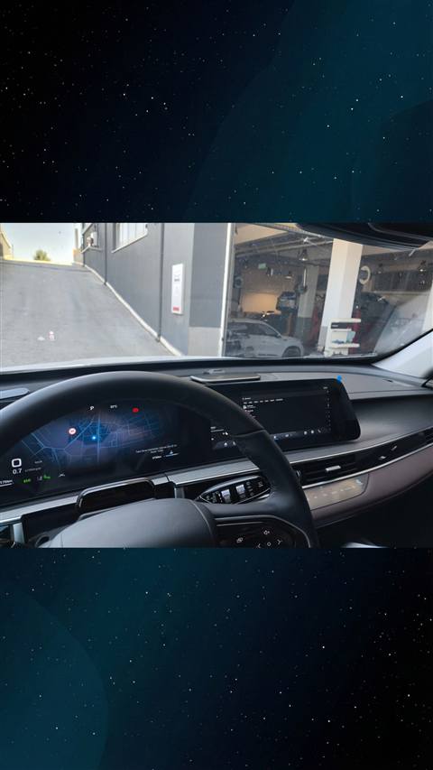 Bir aracin icinden dashboard, direksiyon, dijital gosterge paneli ve navigasyon ekrani gorunmekte, on camdan bir sokak ve servis binasi gorulmektedir.