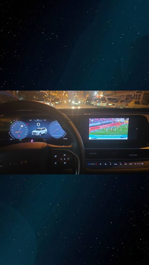 Gece sehir trafiginde bir Chery Tiggo aracinin dijital gosterge paneli ve multimedya ekraninda futbol maci yayini gorunmektedir.