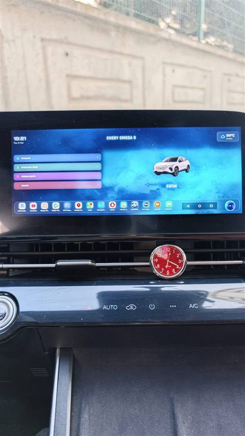 Chery Omoda 5 aracina ait multimedya ekraninda ana menu ve tema arayuzu goruntuleniyor, on konsolda kirmizi bir saat aksesuari bulunuyor.