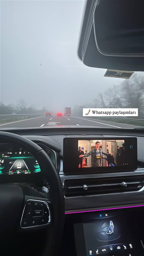 Sisli havada otoyolda ilerleyen bir aracın içinden ön cam, gösterge paneli ve multimedya ekranında video izlenirken çekilmiş bir görüntü.
